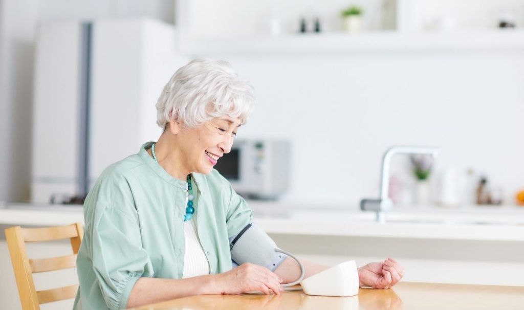Older woman in light blu shirt sitting at table taking a blood pressure reading