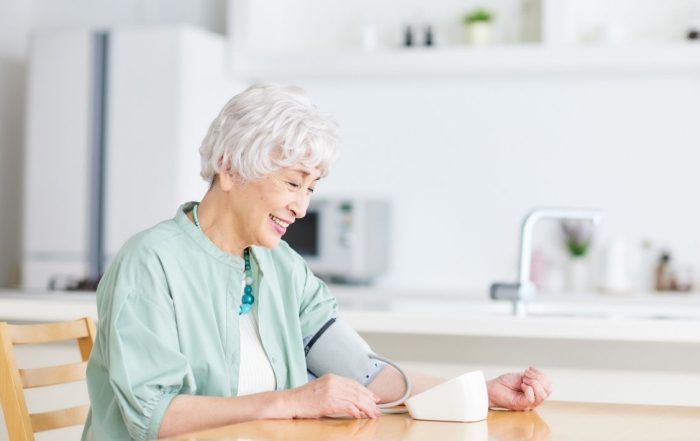 Older woman in light blu shirt sitting at table taking a blood pressure reading
